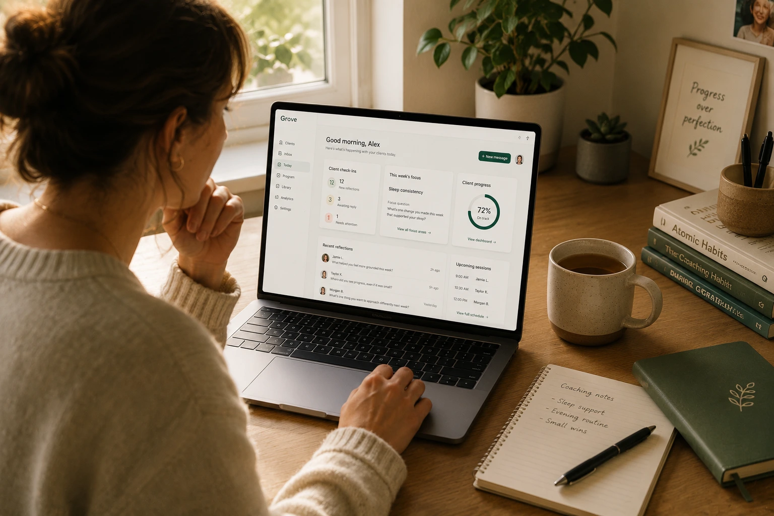 A coach at her desk in morning light, looking over her shoulder at the Grove dashboard on her laptop, with a coffee mug, a small plant, and a notebook on the desk.
